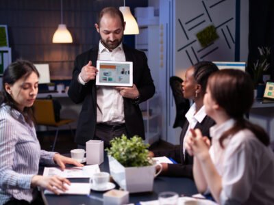 Focused businessman showing corporate graphs presentation using tablet working at company ideas. Diverse multi-ethnic businesspeople brainstorming strategy late at night in office meeting room